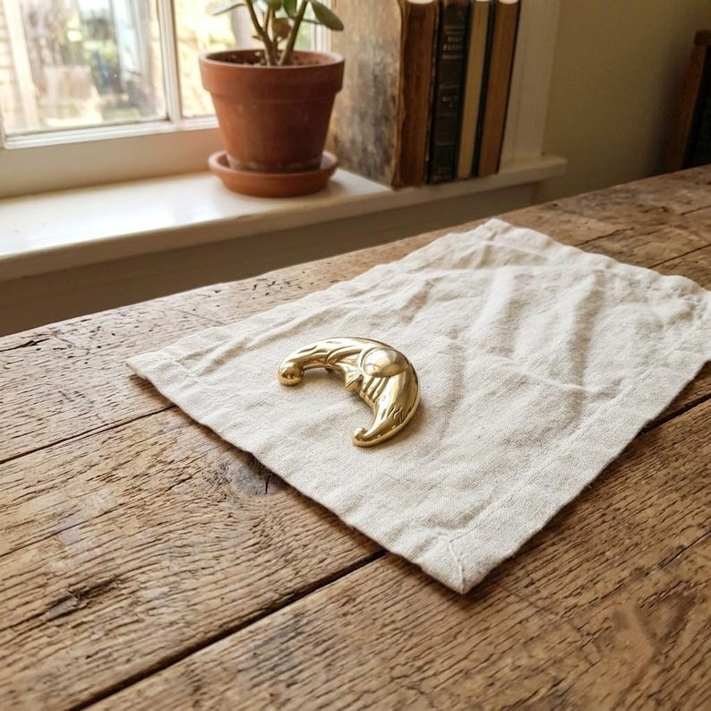 May include: A polished gold-tone metal brooch shaped like a crescent moon with a detailed face. The moon features a curved profile, a smooth surface, and a distinct eye and mouth design. The object is resting on a square piece of off-white fabric.