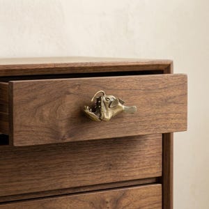 May include: A brass-toned metal drawer pull shaped like an anglerfish with a prominent lure, sharp teeth, and fins. The hardware is mounted on the front of a horizontal wooden drawer, which is part of a larger wooden cabinet with a visible grain.