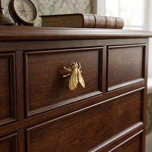 Peut inclure: Une poignée de tiroir en forme d'abeille dorée montée sur une commode en bois brun foncé. L'abeille présente des ailes détaillées et un corps texturé, servant de poignée fonctionnelle pour le tiroir. La commode a un design à cadre.