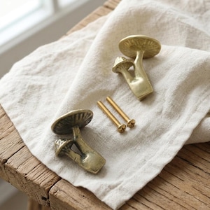 May include: Two brass-coloured metal cabinet knobs shaped like mushrooms with detailed caps and stems, accompanied by two matching gold-toned threaded mounting screws. The knobs feature a smooth, metallic finish and a realistic, organic mushroom silhouette.