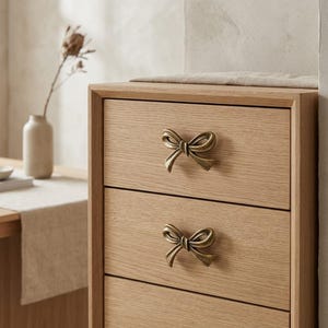 May include: A light-coloured wooden chest of drawers featuring three horizontal drawers. Each drawer is equipped with a decorative, metallic, antique-brass-toned bow-shaped handle centred on the front panel of the drawer.