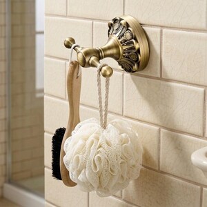 May include: An antique brass-finished wall hook with ornate detailing, featuring two rounded prongs. A wooden-handled brush with black bristles and a white mesh bath sponge are hanging from the hooks, mounted on a light-coloured tiled wall.