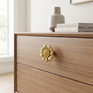 Peut inclure: Un bouton de tiroir en métal doré en forme de tournesol avec un centre texturé et des pétales rayonnants, monté sur une façade de tiroir en bois brun clair. Le bouton présente une finition métallique et un motif floral détaillé en surface.