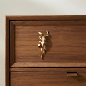 May include: A gold-toned metal lizard-shaped drawer pull mounted on a dark brown wooden drawer front. The lizard figure features a textured surface and is positioned vertically in the center of the rectangular wooden panel with a frame border.