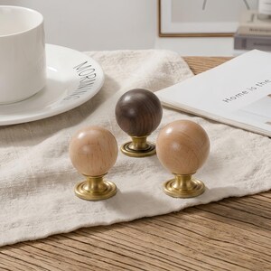 May include: Three spherical cabinet knobs with smooth wooden tops and brushed brass metal bases. One knob features a dark brown wood grain, while the other two knobs are made of a lighter, natural-toned wood. Each knob has a circular metal base.