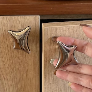 May include: Two polished, silver-toned metal cabinet knobs with a four-pointed, star-like shape. The knobs feature a smooth, reflective surface with curved edges and are mounted on light-colored, wood-grain cabinet doors with vertical texture.