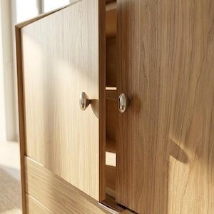 May include: A light brown wooden cabinet featuring two doors with a visible wood grain pattern. Each door is equipped with a small, oval-shaped, metallic silver knob. One door is slightly ajar, revealing an empty interior shelf.