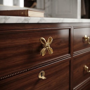 May include: A dark wood dresser featuring a gold-toned metal bow-shaped drawer pull and a matching gold-toned keyhole escutcheon. The drawer front is accented with a beaded trim detail, and a brown book rests on the white marble top surface.