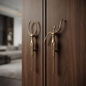 Puede incluir: Dos tiradores de armario de metal dorado montados en puertas de madera de color marrón oscuro. Cada tirador tiene forma de figura humana estilizada con extremidades alargadas, una con los brazos levantados y otra con los brazos extendidos.