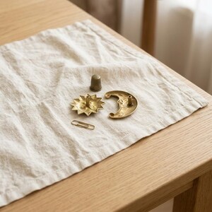 May include: A brass thimble, a star-shaped cabinet knob, a crescent moon-shaped cabinet knob, and a single gold-toned metal paperclip. The items are arranged on a textured, off-white fabric surface, showcasing their metallic finish and details.