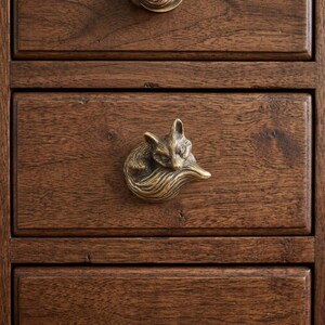 May include: A brass drawer knob shaped like a curled-up fox with detailed fur texture, mounted on a dark brown wooden drawer front. The fox is depicted in a sleeping position with its tail wrapped around its body, featuring a metallic finish.