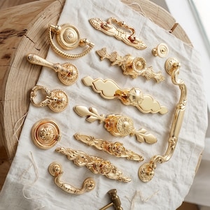 May include: A collection of gold-toned metal cabinet hardware including various drawer pulls, knobs, and drop handles with intricate embossed patterns, floral motifs, and smooth polished surfaces arranged on a white fabric cloth.