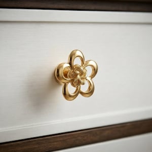 以下が含まれることがあります： 5枚の花びらを持つ花をかたどったゴールドトーンの金属製引き出しノブ。花の中心部には渦を巻くような結び目のデザインが施され、花びらは滑らかなオープンループ状になっています。ノブは白い引き出しの前面に取り付けられています。