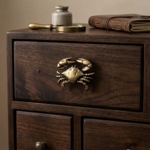 May include: A dark brown wooden drawer featuring a brass-toned metal crab-shaped handle. The crab is depicted with its claws raised and legs extended. The drawer is part of a larger wooden cabinet structure with visible wood grain patterns.