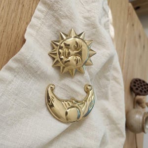 May include: A polished gold-toned metal sun emblem with a central face and radiating triangular points, paired with a matching crescent moon emblem featuring a smooth, curved surface and engraved details on a textured off-white fabric.