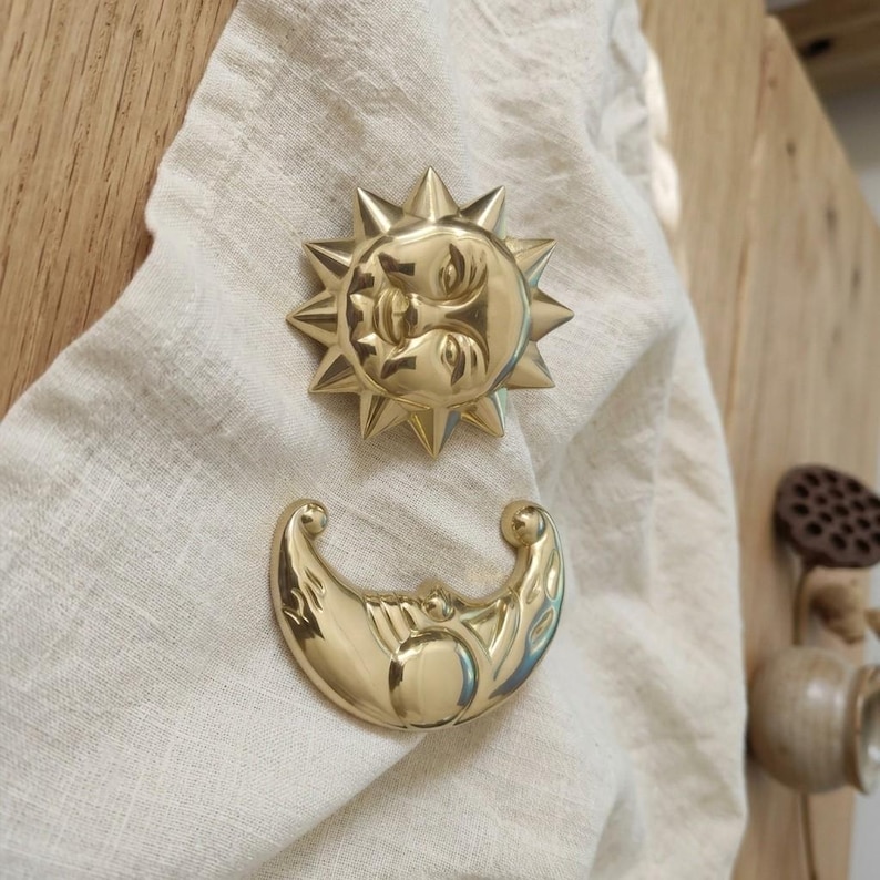 May include: A polished gold-toned metal sun emblem with a central face and radiating triangular points, paired with a matching crescent moon emblem featuring a smooth, curved surface and engraved details on a textured off-white fabric.