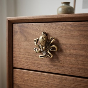 May include: A brass-toned metal cabinet knob shaped like an octopus with textured details on its head and curved tentacles, mounted on a smooth, dark-grained wooden drawer front with a horizontal grain pattern and a visible edge frame.