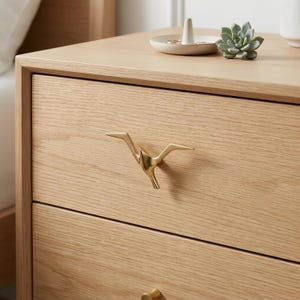 May include: A light-colored wooden nightstand featuring a gold-toned metal drawer pull shaped like a bird in flight. The drawer pull is mounted on the top drawer, which has a visible wood grain pattern. A small white ring dish sits above.