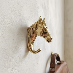 May include: A polished gold-toned metal wall hook shaped like a horse head. The horse head faces to the right, with the neck and mane forming the curved hook structure. The metal surface is reflective and smooth, showing detailed facial features.