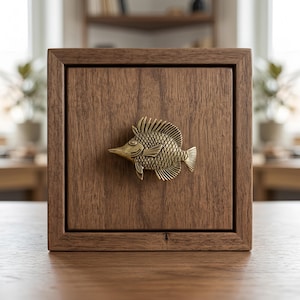 May include: A square wooden box with a dark brown grain finish featuring a centred brass-coloured metal fish ornament. The fish has detailed scales, fins, and a pointed snout, serving as a decorative pull or accent on the front panel of the box.