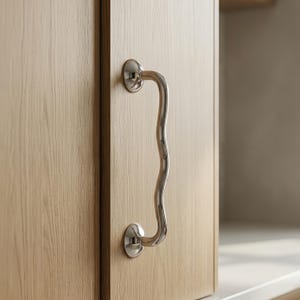 May include: A polished silver-toned metal cabinet handle with a distinct wavy, serpentine shape. The handle is mounted vertically onto a light-colored wooden cabinet door with a visible grain, featuring two circular base plates.