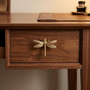 May include: A brass dragonfly-shaped drawer pull mounted on a dark brown wooden drawer. The dragonfly has detailed wings and a segmented body, serving as a functional handle for the furniture piece, which features a natural wood grain finish.
