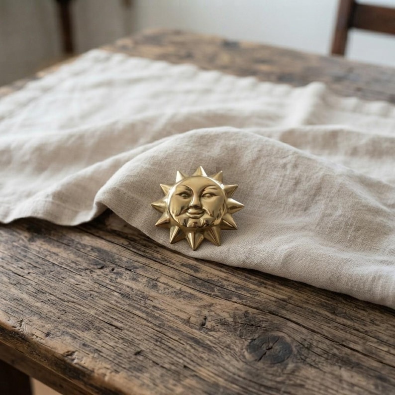 May include: A polished gold-tone metal sun face ornament featuring a central circular face with closed eyes, a small nose, and a smiling mouth, surrounded by a series of triangular rays extending outward in a starburst pattern.