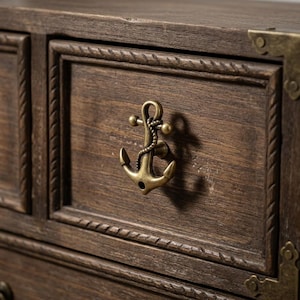 Può includere: Un cassetto in legno marrone scuro con una maniglia in metallo color ottone a forma di ancora nautica con dettagli a corda. Il frontale del cassetto è incorniciato da un bordo intagliato a corda e include una staffa angolare decorativa in ottone.