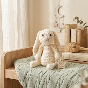 Puede incluir: Un peluche de conejo tejido a ganchillo de color crema con orejas largas y ojos oscuros. Una pequeña etiqueta de madera en forma de estrella unida al brazo del conejo presenta el texto grabado "BABY'S FIRST FRIEND" en una fuente clara.