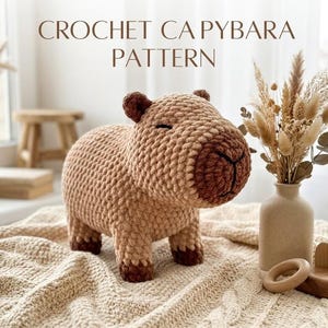May include: A light brown and dark brown crocheted capybara toy with a rounded body, small ears, and a stitched nose. The text CROCHET CAPYBARA PATTERN is displayed above the toy. The toy features a textured yarn surface and four short legs.