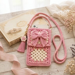 May include: A pink and cream crochet crossbody phone bag featuring a granny square pattern, a flap closure adorned with a large matching crochet bow, and a long strap. A small flower charm with a pink tassel is attached to the side hardware.