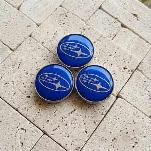 May include: Three circular emblems featuring a vibrant blue background with a silver-toned Subaru logo consisting of an oval outline and six stars of varying sizes. Each emblem has a smooth, glossy finish and a metallic silver border.