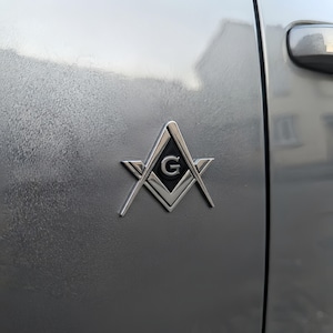 Könnte beinhalten: Ein silberfarbenes Metall-Emblem mit dem freimaurerischen Winkel- und Zirkelsymbol sowie einer zentralen schwarzen Einlage mit dem Buchstaben G, angebracht auf der glatten, metallisch silbernen Oberfläche einer Fahrzeugtür.