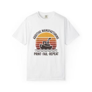 Pode incluir: Uma t-shirt branca de manga curta com um gráfico de um pequeno barco sobre um fundo de pôr do sol. O texto ADDITIVE MANUFACTURING aparece acima do barco e PRINT FAIL REPEAT abaixo. O design apresenta um aspeto desgastado e vintage.
