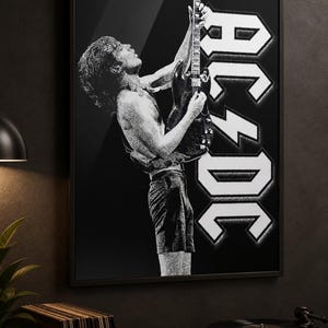 May include: A black and white framed wall art print featuring a side profile of a guitarist playing an electric guitar while looking upward. The iconic AC/DC logo is displayed in large white lettering on the right side of the rectangular composition.