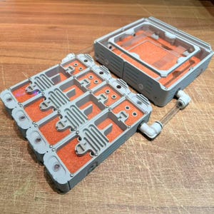 Peut inclure: Un système de fourmilière modulaire composé d'une structure en plastique gris à plusieurs chambres avec un substrat orange et un boîtier carré séparé relié par un tube en plastique transparent pour l'observation.
