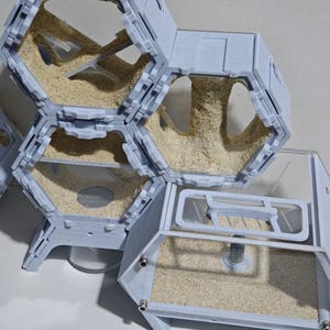 May include: A modular ant farm system featuring interconnected hexagonal and rectangular chambers constructed from light blue speckled plastic. The enclosures contain beige sand substrate, internal structural supports, and clear panels for viewing the interior spaces.
