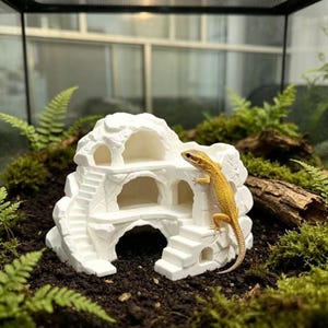 Puede incluir: Un refugio para reptiles en forma de roca blanca de varios niveles con aberturas arqueadas y escaleras integradas. Un dragón barbudo amarillo y naranja está posicionado en el lateral de la estructura, situada en un terrario con tierra y helechos.