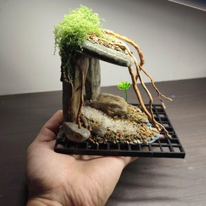 Puede incluir: Un diorama de paisaje en miniatura con pilares de piedra gris que sostienen un techo de piedra plana, adornado con musgo verde brillante y ramas finas de color marrón. La base incluye guijarros, arena blanca y una pequeña planta verde.