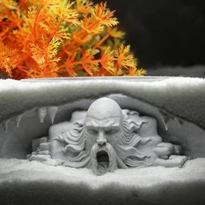 Könnte beinhalten: Eine hellgraue 3D-gedruckte Skulptur, die ein detailliertes Gesicht mit langem, fließendem Bart und Haar zeigt, das aus einer höhlenartigen Struktur hervortritt. Die Figur hat einen offenen Mund und ist von felsigen Formationen mit Stalaktiten umgeben.