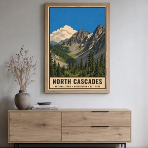 Op de afbeelding: Een verticale poster met een landschapsillustratie van het North Cascades National Park in Washington, opgericht in 1968. Het kunstwerk toont grillige bergtoppen, een diepe vallei en groenblijvende bomen onder een blauwe lucht.