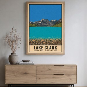 Pode incluir: Um cartaz vertical emoldurado com uma ilustra&ccedil;&atilde;o da paisagem do Parque Nacional Lake Clark, no Alasca. A obra exibe montanhas cobertas de neve, um c&eacute;u azul brilhante, um lago turquesa e um primeiro plano rochoso com o texto LAKE CLARK.