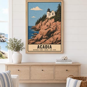 May include: A framed vertical art print featuring a lighthouse perched on rocky cliffs above a blue ocean. The text reads ACADIA NATIONAL PARK MAINE EST. 1919. The illustration includes evergreen trees, a light blue sky with clouds, and a light tan border.