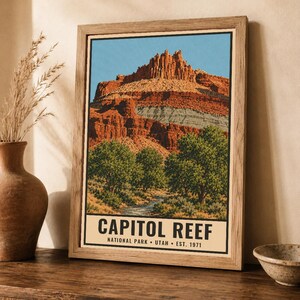 May include: A framed poster featuring a landscape illustration of Capitol Reef National Park in Utah. The artwork displays red rock cliffs, green trees, and a stream under a blue sky. The text reads CAPITOL REEF NATIONAL PARK UTAH EST. 1971.