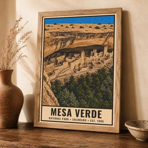 Pu&ograve; includere: Una stampa incorniciata con un'illustrazione paesaggistica del Mesa Verde National Park in Colorado. L'opera raffigura abitazioni rupestri sotto una sporgenza rocciosa, con un cielo blu brillante e alberi verdi. Il testo recita MESA VERDE.