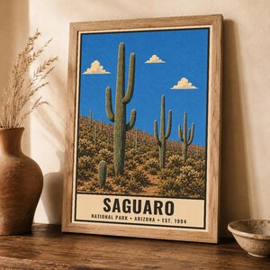 Op de afbeelding: Een ingelijste wandkunstprint met een woestijnlandschap met hoge groene saguaro-cactussen onder een helderblauwe lucht met drie kleine wolken. Het onderste gedeelte bevat de tekst SAGUARO NATIONAL PARK ARIZONA EST. 1994 in zwarte letters.