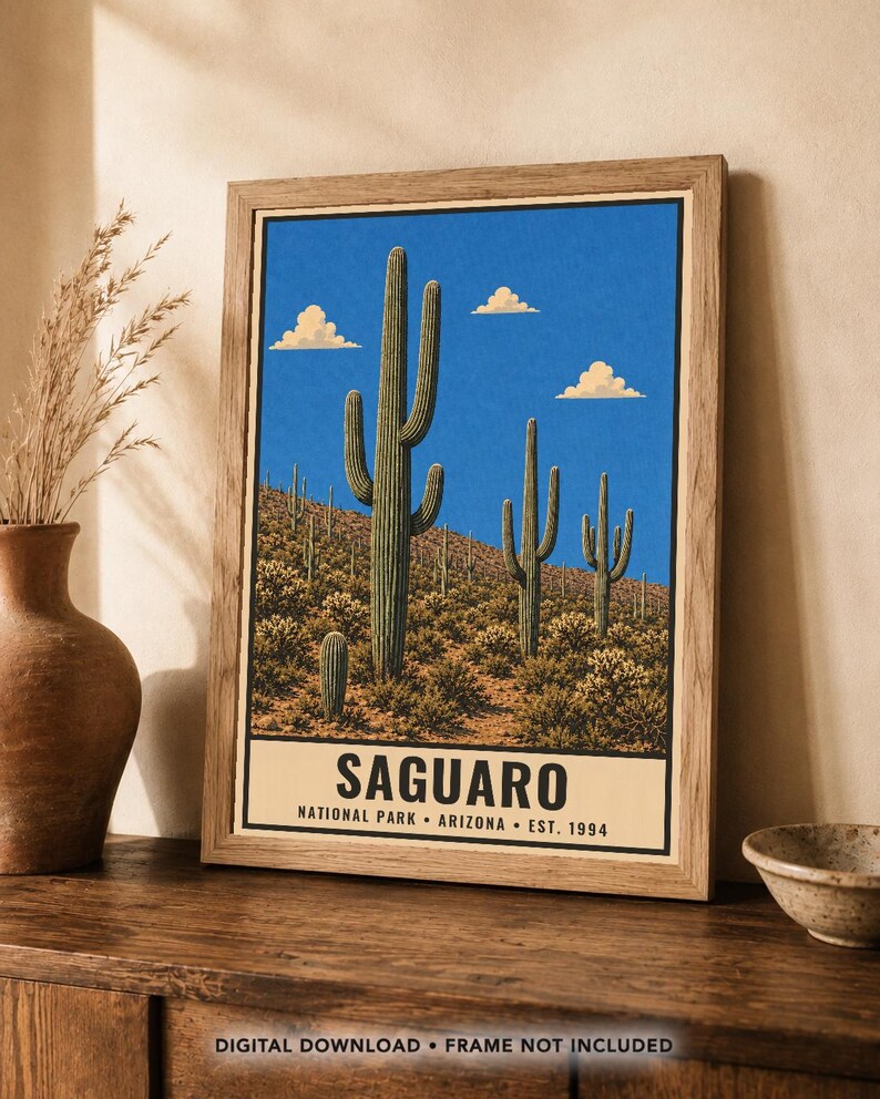 Op de afbeelding: Een ingelijste wandkunstprint met een woestijnlandschap met hoge groene saguaro-cactussen onder een helderblauwe lucht met drie kleine wolken. Het onderste gedeelte bevat de tekst SAGUARO NATIONAL PARK ARIZONA EST. 1994 in zwarte letters.