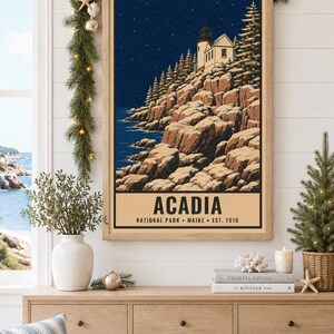 Puede incluir: Un cuadro rectangular enmarcado que muestra una escena costera nocturna con un faro sobre un acantilado rocoso y &aacute;rboles de hoja perenne bajo un cielo estrellado. El texto en la parte inferior dice ACADIA NATIONAL PARK MAINE EST. 1919 en negro.