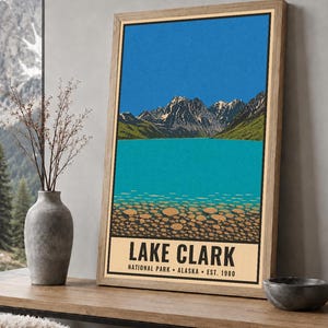 Pode incluir: Um cartaz vertical emoldurado com uma ilustra&ccedil;&atilde;o estilizada do Parque Nacional Lake Clark, no Alasca. A obra retrata um c&eacute;u azul brilhante, montanhas escarpadas com neve, um lago turquesa e um primeiro plano rochoso com o texto LAKE CLARK.