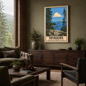 Op de afbeelding: Een ingelijste verticale poster met een landschapsillustratie van Voyageurs National Park in Minnesota. Het kunstwerk toont een blauw meer met rotsachtige eilanden, hoge dennenbomen op de voorgrond en een wolk in een blauwe lucht.